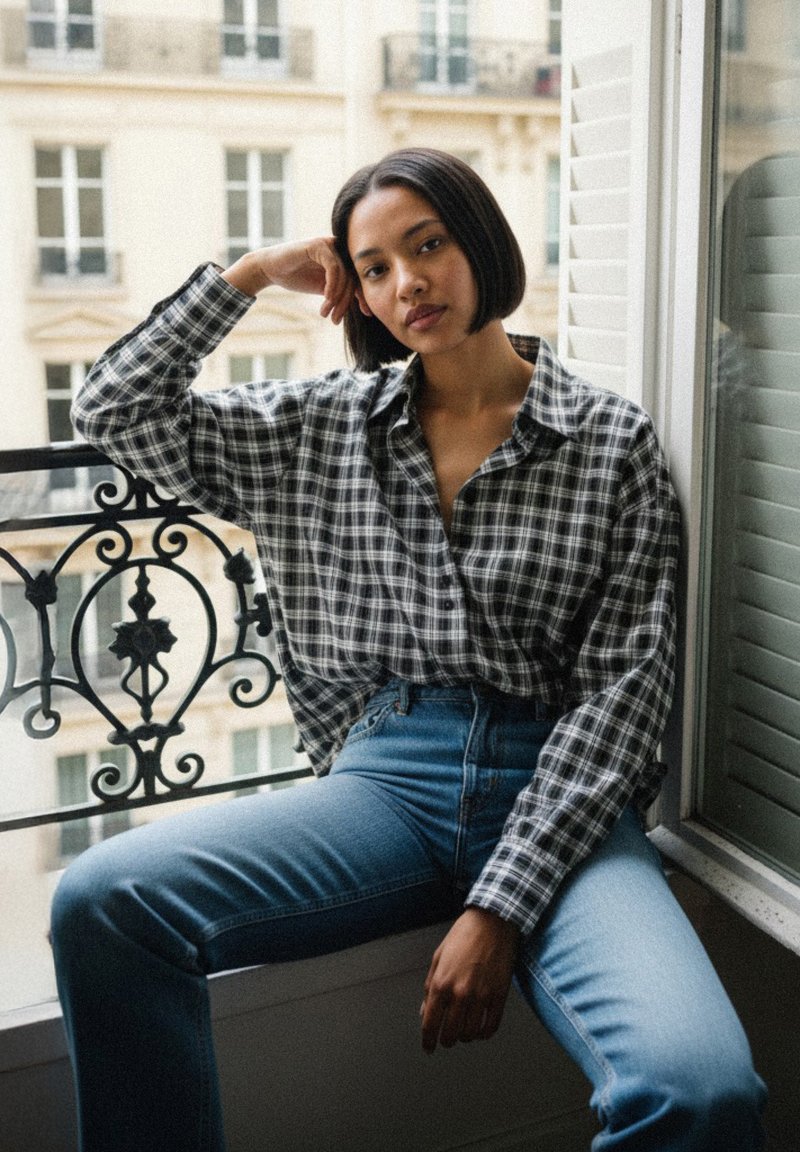 Black and white checkered button-up shirt paired with high-waisted blue jeans. Sitting by a window with decorative black iron railing.