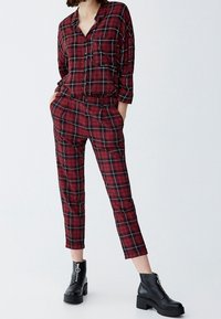 Ensemble à carreaux en rouge, noir et blanc. Chemise boutonnée à manches longues et pantalon avec poches latérales. Porté avec des bottes noires à la cheville.