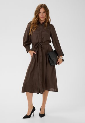 Femme en robe marron à manches longues boutonnée avec une taille nouée, tenant une pochette noire, portant des talons hauts noirs, debout sur un fond uni.