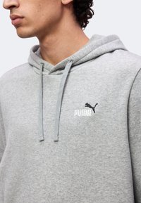 Άνδρας με ανοιχτό γκρι φούτερ Puma με κορδόνια και το λογότυπο Puma στο στήθος, στραμμένος προς τα αριστερά σε απλό φόντο.