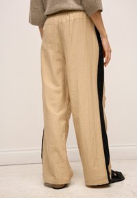Beige vidde bukser med en tekstureret overflade, prydet med sorte sidestriber. Elastisk talje og afslappet pasform, vist fra bagsiden.