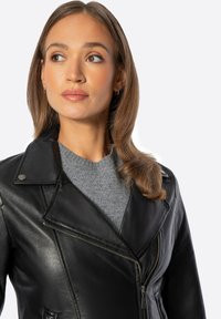 Chaqueta de motociclista de cuero negro con solapa, cremallera frontal y herrajes plateados sobre un suéter gris. Textura suave y diseño ajustado.