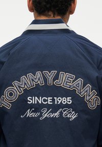 Άτομο που φοράει μπλε σκούρο μπουφάν με κεντημένο στο πίσω μέρος το κείμενο "TOMMY JEANS SINCE 1985 New York City", στραμμένο με την πλάτη προς την κάμερα.