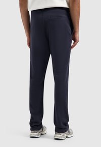 Marineblauwe sweatpants met een gladde textuur, voorzien van een achterzak en een elastische tailleband, gecombineerd met lichtgrijze hardloopschoenen.