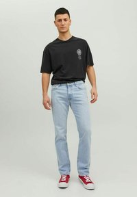 Camiseta negra de gran tamaño con detalle gráfico, jeans rectos de mezclilla azul claro y zapatillas rojas con suelas blancas, de pie contra un fondo neutro.