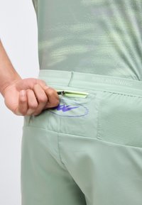 Χέρι που τραβάει το φερμουάρ της μικρής πίσω τσέπης σε ανοιχτό πράσινο σορτς Nike Running, υπογραμμίζοντας την υφή του υφάσματος και τη σήμανση.