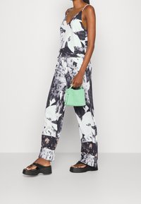 Jumpsuit con motivo floreale in bianco e nero con spalline sottili, abbinato a una borsa verde. Presenta pantaloni a gamba larga e sandali neri chunky.