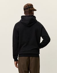 Felpa nera con cappuccio e tasca a marsupio, con polsini e orlo a coste, indossata con pantaloni a quadri marroni e un berretto a scacchi.