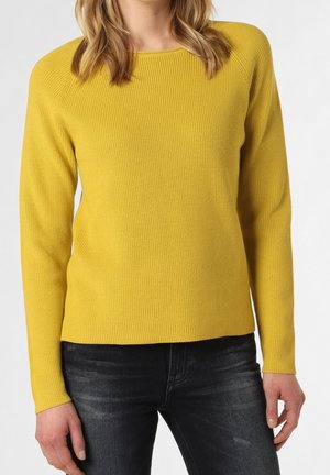 Vrouw draagt een mosterdgele geribbelde gebreide trui met lange mouwen en zwart vervaagde jeans, staand tegen een effen achtergrond.