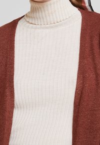 Ribbed-tekstuurinen kermaisenvärinen pooloneule kerrostettuna rustinvärisen teksturoidun neulepaidan alle, korostaen pehmeitä materiaaleja ja rentoa istuvuutta.