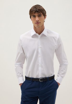 Ein Mann trägt ein eng anliegendes weißes Hemd mit Knöpfen, das in marineblaue Hosen mit einem schwarzen Gürtel gesteckt ist, und steht mit den Händen in den Taschen vor einem schlichten Hintergrund.