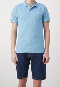 Jasnoniebieska koszulka polo z białymi akcentami przy kołnierzyku i rękawach, wykonana z miękkiej bawełny, w połączeniu z ciemnoniebieskimi szortami dżinsowymi.