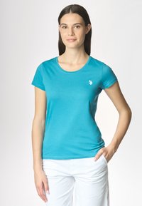 Una donna con lunghe capelli castani indossa una maglietta turchese e pantaloni bianchi, in piedi con sicurezza con una mano sulla vita e un leggero sorriso.