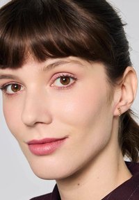 Złote kolczyki o wiszącym wzorze, z małymi koralikami. Widoczna gładka skóra, lekki makijaż i brązowe włosy z grzywką.