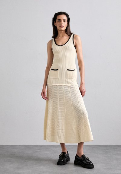 Conjunto de dos piezas de punto que incluye una blusa color crema con borde negro y bolsillos, combinada con una falda de crema fluida y texturizada. Unos zapatos negros gruesos completan el look.