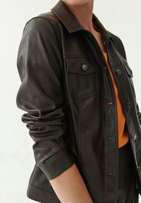 Giacca in pelle nera con colletto strutturato, due tasche frontali e chiusura con bottoni. Le maniche sono leggermente arricciate ai polsini.