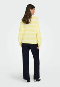 Gelb-weiß gestreifter Strickpullover mit langen Ärmeln, lockerer Passform, geripptem Saum, getragen zu dunklen, weit geschnittenen Hosen. Strukturierter Stoff.