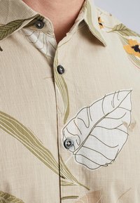 Kortærmet beige skjorte med lodrette striber og blomsterdesign i grøn og hvid, med mørke knapper ved kraven.