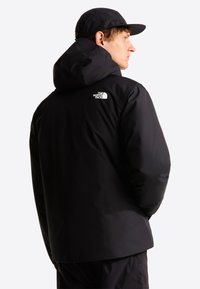 Άνδρας που φοράει μαύρο μπουφάν The North Face με κουκούλα και μαύρο καπέλο που ταιριάζει, φαίνεται από πίσω σε απλό φόντο.