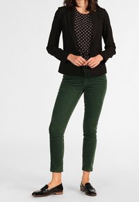 Vrouw in een zwart colbert, met een geblokte zwarte top, groene broek en zwarte instappers, staand met de handen voor zich gekruist.