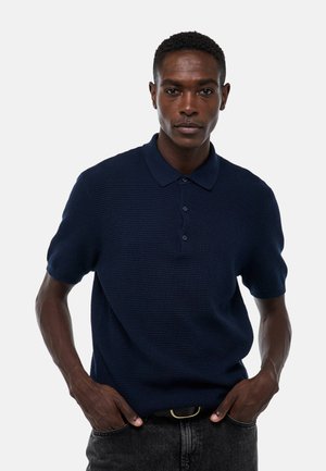 Polo shirt bleu marine à manches courtes avec un motif en maille texturé, patte de boutonnage à trois boutons et col classique. Porté avec un jean foncé et une ceinture noire.