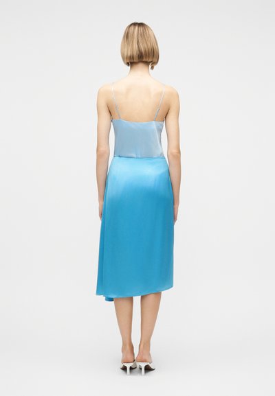 Γυναίκα με κοντά ξανθά μαλλιά, φοράει ανοιχτό μπλε αμάνικη μπλούζα και μπλε μίντι φούστα που ταιριάζει, στέκεται ξυπόλυτη σε λευκές μπαλαρίνες, από πίσω.