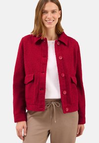Femme portant une veste rouge à boutons texturée sur une chemise blanche et un pantalon beige à cordon, debout et souriante sur un fond uni.