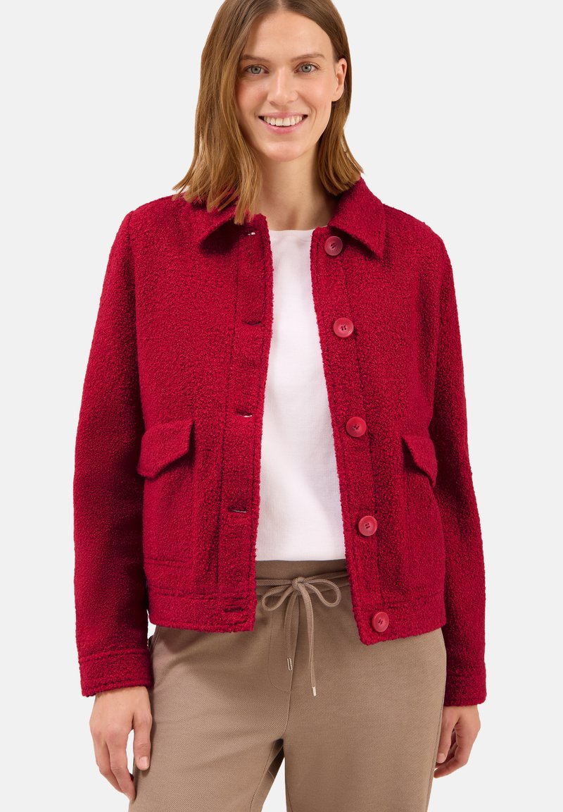 Femme portant une veste rouge à boutons texturée sur une chemise blanche et un pantalon beige à cordon, debout et souriante sur un fond uni.