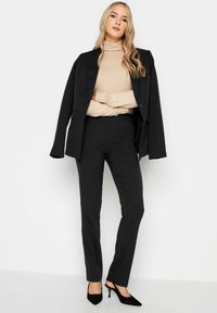 Svarta kavajer och byxor med en beige polotröja. Kavajen har en enda knapp och en smidig silhuett; byxorna är skräddarsydda med en lätt glans.