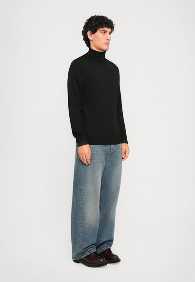 Mann steht vor weißem Hintergrund und trägt einen schwarzen Rollkragenpullover, ausgebleichte weit geschnittene Jeans und dunkelbraune Schuhe, mit Blick nach rechts.