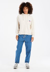 Felpa beige con cappuccio e cordino, tasca frontale e piccolo logo. Abbinate a pantaloni di denim blu larghi e sneakers grigie.