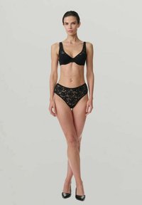 Femme debout en soutien-gorge en dentelle noire et culotte taille haute, portant des chaussures à talons noirs, sur un fond gris uni.