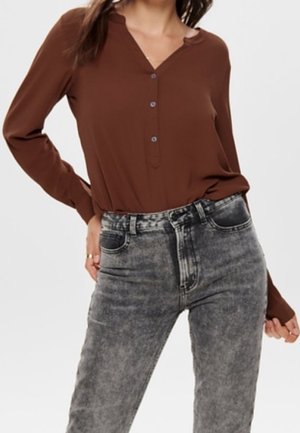 Femme portant une chemise marron à manches longues boutonnée rentrée dans un jean gris délavé taille haute sur un fond uni.