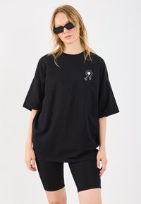 T-shirt oversized preto em algodão com uma gráfica de flor branca no peito esquerdo, combinado com calções de bicicleta pretos com relevos.