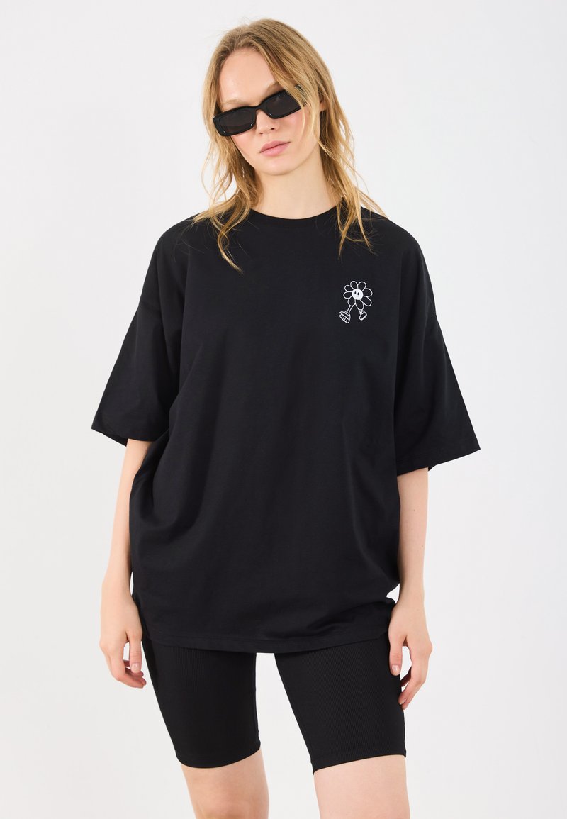 T-shirt oversized preto em algodão com uma gráfica de flor branca no peito esquerdo, combinado com calções de bicicleta pretos com relevos.