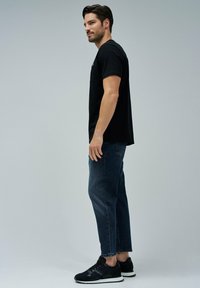Hombre de pie de lado, vistiendo una camiseta negra, jeans oscuros y zapatillas negras frente a un fondo gris claro liso.