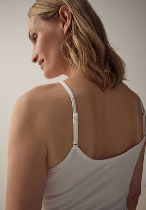 Femeie cu păr blond până la umeri, purtând un top alb cu bretele subțiri, văzută din spate și uitându-se în lateral.