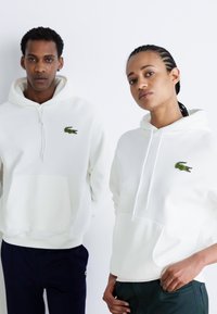 Fehér kapucnis pulóverek kenguru zsebbel, zöld Lacoste krokodil logókkal. Sima felület, zsinórok a kapucniban; sötét nadrággal viselve.