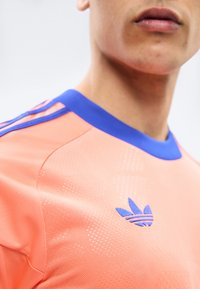 Gros plan d'une personne portant un t-shirt de sport corail avec un col bleu, un logo Adidas bleu, et des bandes bleues sur l'épaule.