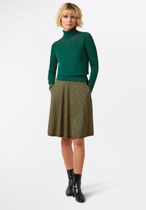 Grüner Strickrollkragensweater mit strukturierten Details, kombiniert mit einem plissierten Rock mit einem geometrischen Schwarz-Gold-Muster und Taschen.
