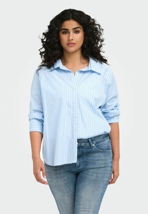 Vrouw met krullend donker haar, die een lichtblauw-wit gestreept overhemd met knopen en een blauwe spijkerbroek draagt tegen een effen achtergrond.