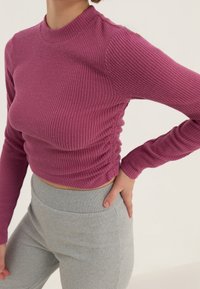 Langärmliges, geripptes Oberteil in Mauve mit Rundhals und seitlicher Raffung, kombiniert mit schlanken, hellgrauen Hosen. Glatte Textur und schmeichelhafte Passform.