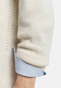 Cream-colored knitted sweater with a textured body and ribbed hem, layered over a light blue collared shirt. Visible seam detailing.