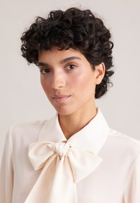 Light cream blouse with a large bow tie at the collar, featuring a smooth texture and soft fabric. Simple design with clean lines.
