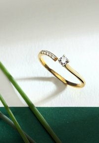 Guldring med en solitär diamant och en böjd band med små klara stenar. Slät yta, minimalistisk design och öppen form.