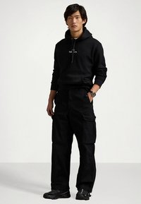 Μαύρο φούτερ με κουκούλα και τσέπη στο μπροστινό μέρος, συνδυασμένο με μαύρο παντελόνι cargo με πλαϊνές τσέπες. Φορεμένο με μαύρα αθλητικά παπούτσια.