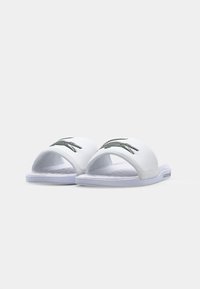 Sandale albe cu talpă deschisă, având o branț texturat și un logo Lacoste verde închis pe curea, accentuate cu o talpă albă.
