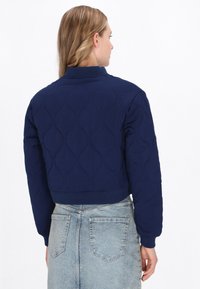Femeie cu păr lung, blond, îmbrăcată într-o jachetă bleumarin matlasată și o fustă din denim de un albastru deschis, întoarsă cu spatele într-un fundal alb simplu.