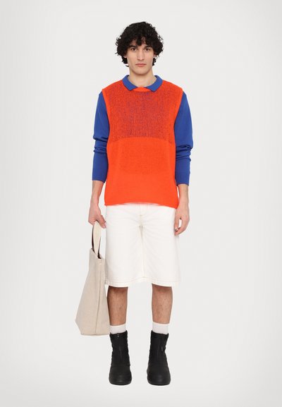 Pullover lavorato a maglia con corpo arancione e maniche blu, abbinato a pantaloncini bianchi e stivali neri alla caviglia. La modella tiene una borsa a tracolla beige.