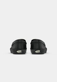 Schwarze Slipper mit strukturiertem Finish, abgerundetem Absatz und weißem Fleece-Futter. Mit einem sichtbaren Logo-Akzent auf der Sohle.